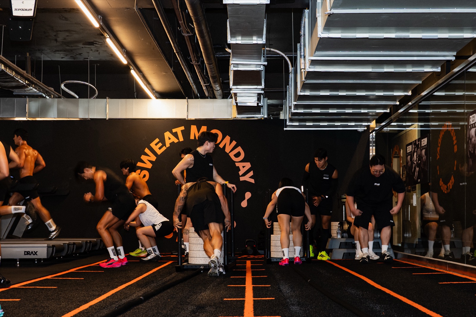 Orange-lined training floor under the SWEAT MONDAY wall graphic — sled push lanes and treadmill runs at the FREEDOM community workout in Seoul